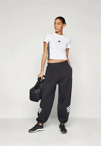 Top atlético cropped branco com logo preto, combinado com calças de treino largas pretas com detalhes em riscas brancas. Sapatos atléticos pretos e uma bolsa.
