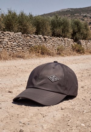 Une casquette de baseball Replay gris foncé à visière courbée repose sur un chemin poussiéreux, près d’un mur en pierres sèches et d’arbustes.