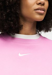 Κοντινό πλάνο ενός ατόμου που φοράει ροζ φούτερ Nike με λευκό λογότυπο Nike, εμφανίζοντας τον λαιμό, τα χείλη και τα σγουρά μαλλιά.