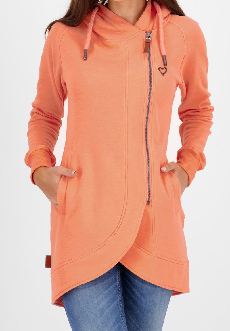 Haut à capuche de couleur pêche avec une fermeture éclair sur le devant, des poches latérales et un ourlet arrondi. Il est doté de cordons de serrage et d'un petit logo en forme de cœur sur la poitrine.