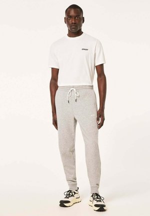 Weißes T-Shirt mit schwarzem Logo, graue Sweatpants mit elastischem Bund und Kordelzug, strukturiertes Material, getragen mit schwarz-weißen Sneakers.