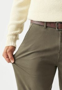 Sile rohelised püksid siledast kangast, sujuva lõikega, mille hoidmiseks on pruun nahkvöö, näidates tekstuurset tikandit ja vormitud taljet.