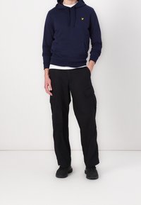 Sudadera con capucha azul marino con un logo amarillo en el pecho, combinada con pantalones cargo negros con bolsillos laterales y un corte holgado, usados con zapatos negros.