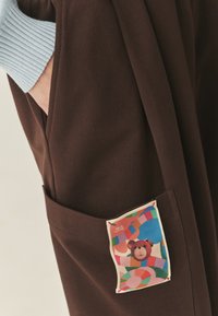 Brązowe bawełniane spodnie z boczną kieszenią, na której znajduje się kolorowa naszywka przedstawiająca niedźwiedzia na abstrakcyjnym tle geometrycznych kształtów.