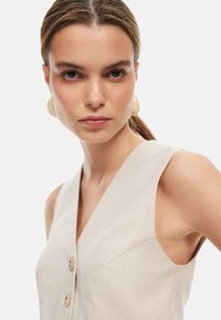 Beige gestreifter ärmelloser Top mit tiefem V-Ausschnitt, der mit zwei Knöpfen und einem subtilen strukturierten Stoff versehen ist. Die Modells trägt große runde Ohrringe.