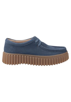 Zapato de cuero azul con punta redondeada, costuras decorativas y una suela de goma marrón gruesa y estriada. Presenta cordones y un acabado suave.
