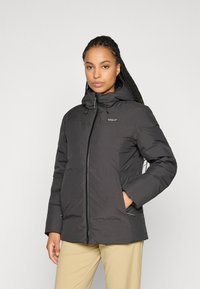 Vrouw met gevlochten haar, gekleed in een zwarte winterjack met capuchon en ritssluiting, en kaki broek, met handen in de zakken van de jas, op een neutrale achtergrond.