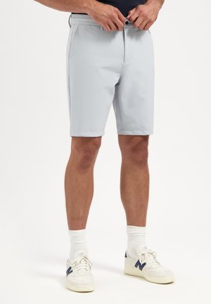 Man fastening button on light gray knee-length shorts, wearing white socks and white sneakers with navy accents.