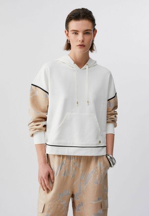 Donna che indossa una felpa con cappuccio bianca con maniche beige a motivi floreali e pantaloni cargo beige coordinati, accessoriate con braccialetti d'argento.