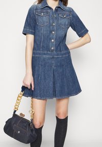 Vestido de mezclilla de manga corta con dos bolsillos en el pecho, cierre de botones en la parte delantera y falda acampanada. Combinado con un bolso azul oscuro con cadena.