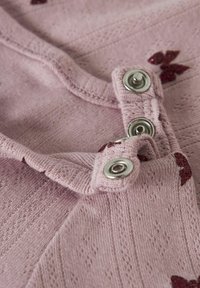 Roza pletenina z teksturiranimi vzorci, opremljena z metalnimi pritisnimi gumbi in dekorativnimi rdečimi bleščicami v obliki metulja.