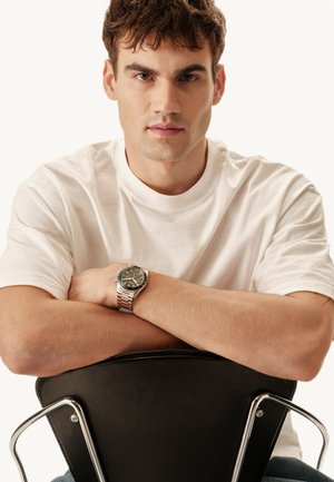 Montre-bracelet en argent avec un cadran noir, bracelet en acier inoxydable, portée au poignet d'une personne portant un t-shirt blanc uni, bras croisés sur une chaise noire.