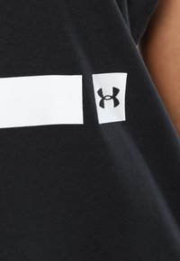 Detailní záběr na černou tkaninu s bílým obdélníkovým designem a malým bílým čtvercem s logem Under Armour v černé barvě.