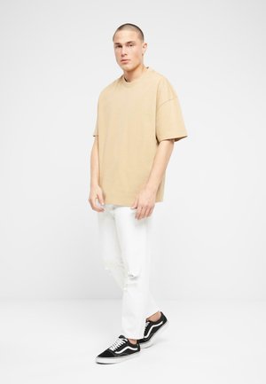 Camiseta de algodón beige de gran tamaño, con corte holgado; combinada con jeans blancos desgastados y zapatillas Vans negras con acentos blancos.