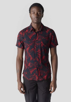 Joven con camisa de manga corta negro y roja con patrones de hojas, de pie frente a un fondo claro y liso.