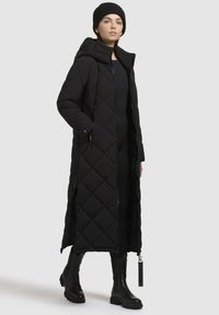 Langer schwarzer Daunenmantel mit Steppmuster, frontalem Reißverschluss und Kapuze. Kombiniert mit einer schwarzen Strickmütze und kniehohen schwarzen Stiefeln.