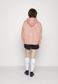 Persona che indossa una felpa rosa con cappuccio con testo circolare bianco "PARIS A R E" sulla schiena, pantaloni corti neri, calze bianche fino al ginocchio e scarpe da calcio nere.