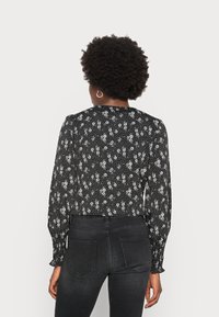 Blusa preta com padrão floral branco, mangas longas com punhos franzidos, decote alto, usada com jeans pretos ajustados.