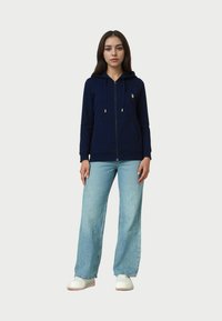 Tengerészkék cipzáras kapucnis pulóver arany színű részletekkel, kenguru zsebekkel, világoskék, bő szárú farmerrel és fehér, slip-on cipőkkel párosítva. Simító anyag, laza fazon.