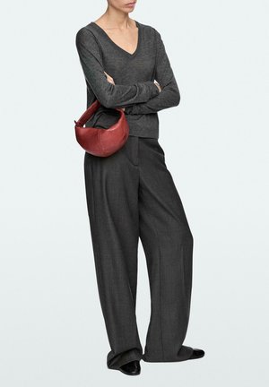 Personne portant un pull col en V gris foncé et un pantalon large, tenant un petit sac à main en cuir rouge sous le bras, debout les bras croisés.