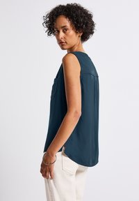 Top senza maniche di colore teal con orlo arrotondato, caratterizzato da una piega sul retro e una tasca singola. Tessuto morbido e leggero.