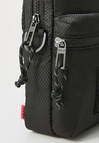 Close-up of black textured backpack corner with two silver zippers labeled “HUGO” and black cord pulls, and a small red fabric tag.