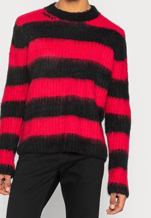 Persoon die een pluizige rood-zwarte trui met brede strepen en lange mouwen draagt, gecombineerd met een zwarte broek, tegen een lichte achtergrond.