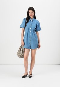 Vestido camiseiro de denim com mangas bufantes curtas, fecho frontal com botões e pregas subtis. Combinado com uma mala de padrão pele de cobra e sapatos pretos de bico fino.