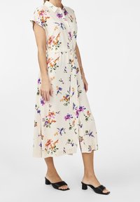 Bloemen midi-jurk met korte mouwen, zachte beige stof, levendige oranje en paarse bloemen, geaccentueerde taille, split aan de zoom, gecombineerd met zwarte high-heeled muiltjes.