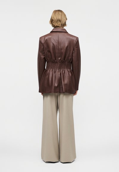 Άτομο με κοντά ξανθά μαλλιά φοράει ένα καφέ δερμάτινο μπουφάν με σφηκοφωλιά στη μέση και μπεζ παντελόνια με φαρδιά μανίκια, στέκεται μπροστά σε απλό φόντο.