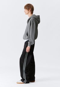 Grauer Kapuzenpullover mit einer vorderen Tasche, kombiniert mit schwarzen Hosen, die ein kontrastierendes Zickzackmuster an der Seite aufweisen, sowie weißen Turnschuhen.