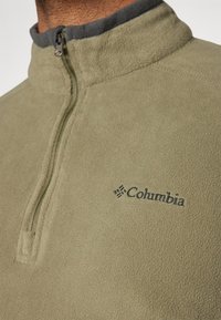 Jasnooliwkowy polarowy pulower z półzamkiem, szarym kołnierzem i małym wyszywanym logo Columbia z przodu. Miękka tekstura.