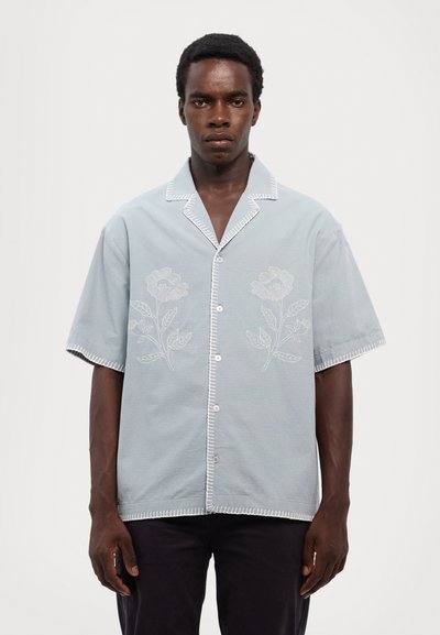 Šviesiai mėlynas trumpomis rankovėmis marškinėliai su išsiuvinėtais gėlių raštais, užsegami mygtukais priekyje, ir kontrastinga balta siūlelių apdaila aplink apykaklę ir pakraštį.