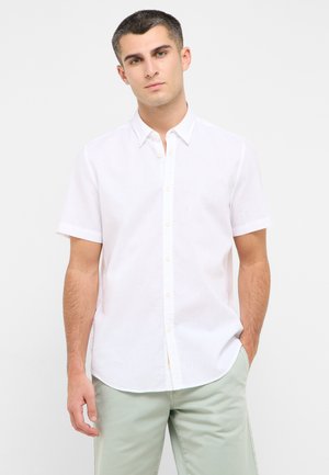 Kurzärmliges, weißes Leinenhemd mit Button-Down-Kragen und sichtbaren Knöpfen; getragen mit grünen Hosen, die eine entspannte Passform zeigen.