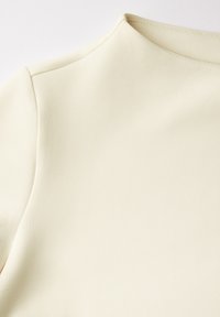 Tessuto color crema, texture liscia, design attillato con un colletto alto, con dettagli di cucitura evidenti lungo la spalla.