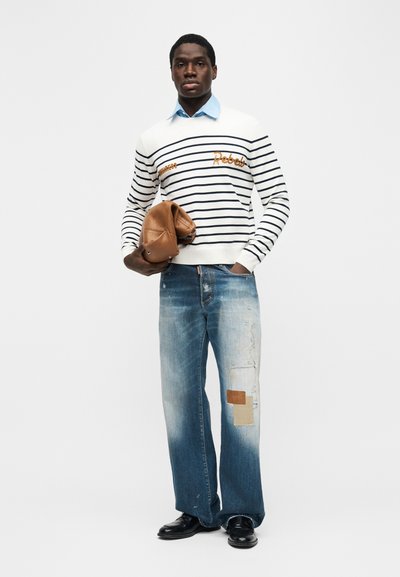 Homme portant un pull à rayures blanches et bleu marine par-dessus une chemise bleue, jean large délavé avec des patchs, chaussures noires, tenant une veste en cuir marron pliée.