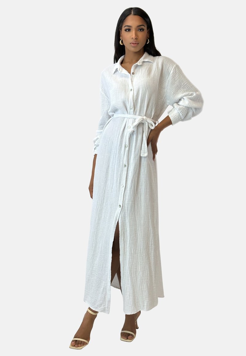 Vestido camisero blanco de manga larga en algodón texturizado, con cierre de botones, cinturón en la cintura y una abertura frontal. Usado con sandalias con tiras.