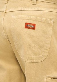 Pantalon beige Dickies avec une poche arrière ornée d'un petit patch logo Dickies rouge et jaune cousu sur la poche.