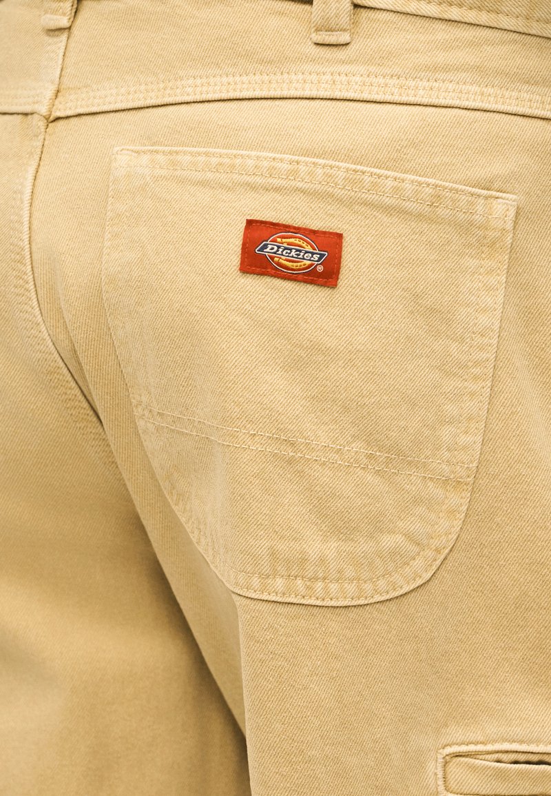 Pantalones beige Dickies con bolsillo trasero que presenta un pequeño parche con el logo de Dickies en rojo y amarillo cosido en el bolsillo.