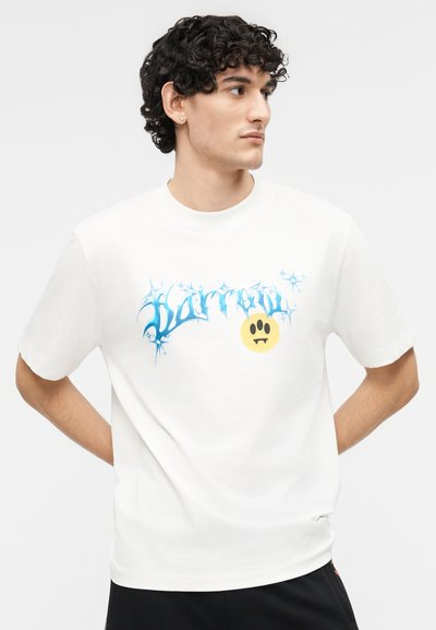 Νέος άνδρας με σγουρά μαλλιά που φοράει λευκό μπλουζάκι με μπλε κείμενο σε στυλ γκράφιτι και κίτρινο γραφικό με το χαμόγελο ενός βρικόλακα.