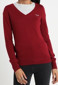 Röd V-ringad tröja i mjukt stickat material, med långa ärmar och ribbade manchetter. Logotypen på vänster bröst. Bärs över en grå skjorta.