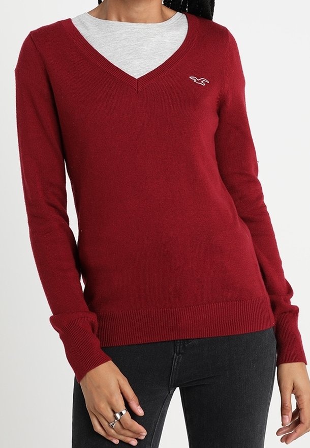 Röd V-ringad tröja i mjukt stickat material, med långa ärmar och ribbade manchetter. Logotypen på vänster bröst. Bärs över en grå skjorta.