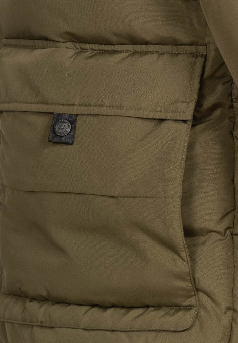 Chaqueta aislante de color verde oliva oscuro con una textura acolchada, que cuenta con un bolsillo frontal y un parche de logo de tela negra en la costura del bolsillo.