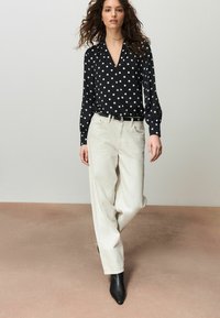 Fekete pöttyös blúz V-nyakkivágással és hosszú ujjakkal, világos bézs bő szárú nadrággal és fekete, hegyes orrú bokacsizmával párosítva.