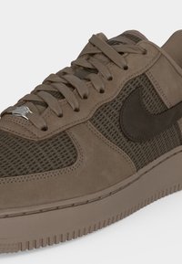 Καφέ αθλητικό παπούτσι Nike Air Force 1 με πάνελ σουέτ και διχτυωτά, κορδόνια που ταιριάζουν, μαύρο λογότυπο Swoosh και μεταλλική ετικέτα κορδονιών σε λευκό φόντο.