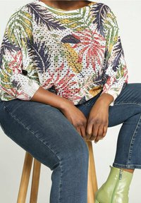 Top de manga larga de punto suelto con un colorido estampado de hojas tropicales, combinado con jeans azul oscuro y botas verdes hasta el tobillo, sentado en un taburete de madera.