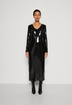 Μαύρο φόρεμα με παγιέτες, μακριά μανίκια και V-λαιμό, με εφαρμοστό σχέδιο και μήκος μίντι, συνδυασμένο με μαύρες μπότες με τακούνι.