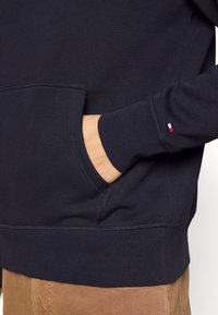 Marineblaues Sweatshirt mit einer Kängurutasche und gerippten Bündchen. Verfügt über ein kleines rot-weiß-blaues Logo-Accessoire am linken Ärmel. Weiche Textur.