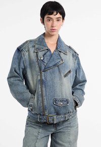 Model met kort zwart haar draagt een blauwe spijkerjack en bijpassende spijkerbroek, staande tegen een effen witte achtergrond.