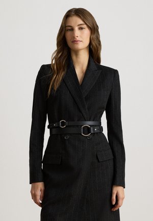 Femme aux longs cheveux bruns porte une robe blazer noire à fines rayures avec une taille ceinturée double et des poches à rabat, debout devant un fond uni.